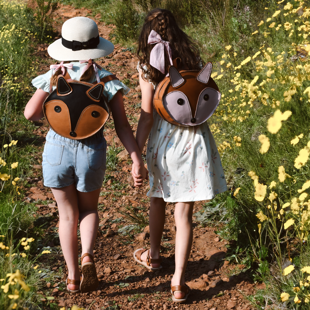 Two children walking outdoors holding hands, wearing fox-shaped backpacks.