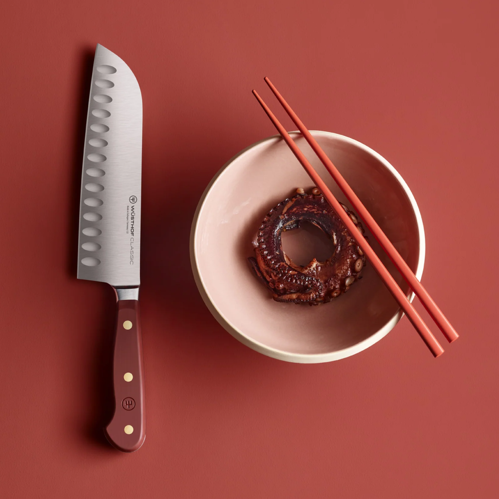 Santoku knife with a red handle next to a bowl of food and chopsticks on a red background