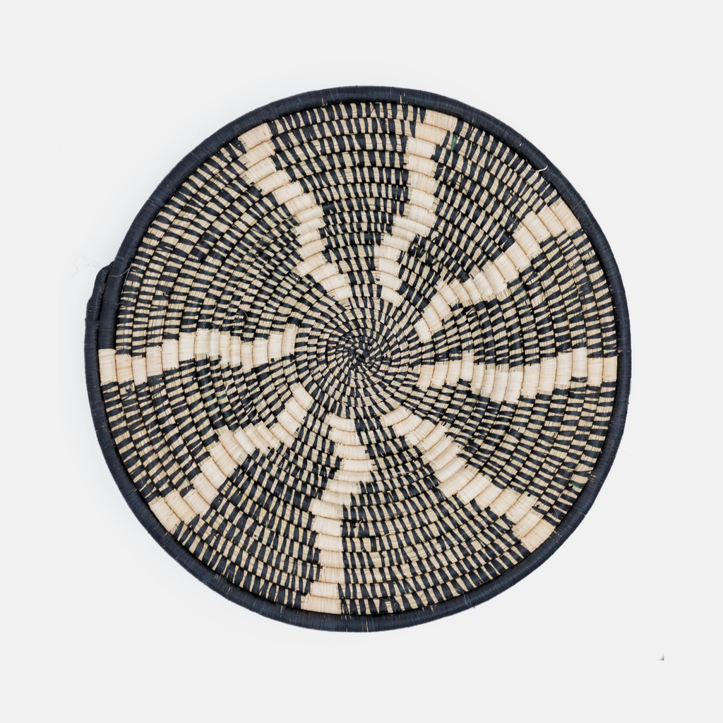 A round woven basket or tray with a black and natural beige spiral pattern, photographed from directly above on a light gray background.