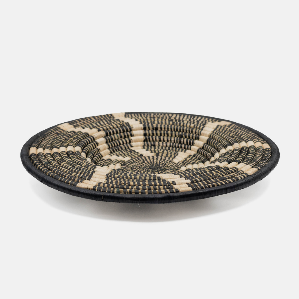 A round woven basket tray with a black and natural beige geometric pattern, photographed on a clean white background.
