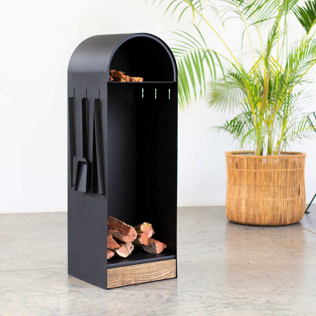 A black firewood holder made of powder-coated steel with a solid oak door, containing firewood and with a tool hanging on the side.