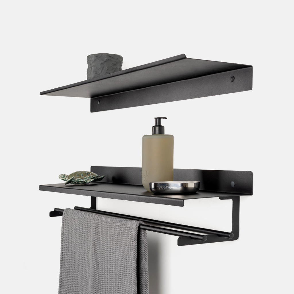 Black metal wall-mounted bathroom shelf set with a towel bar, shown with a gray towel, soap dispenser, small bowl, and decorative item against a light gray background.