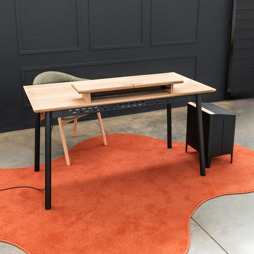 Wooden desk with black legs on a red rug against a dark wall.