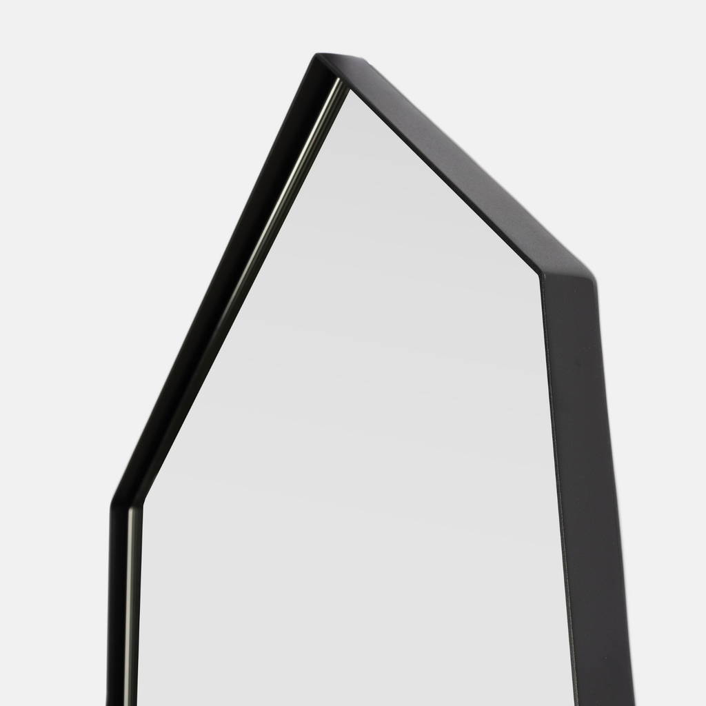 A close-up of a modern hexagonal wall mirror with a matte black frame, showing a light gray reflective surface against a light gray background.