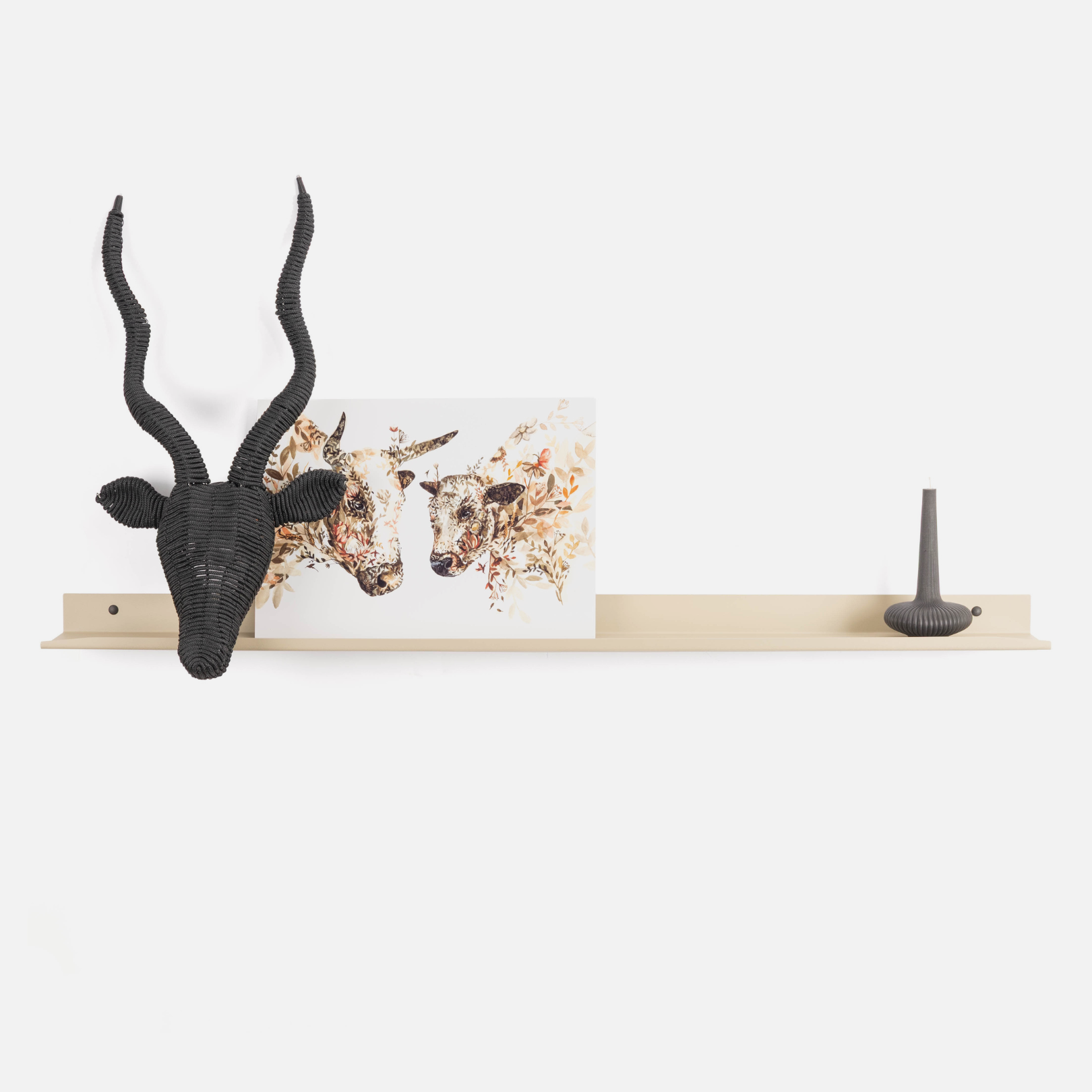A minimalist steel floating shelf mounted on a white wall, styled with a black woven antelope head wall sculpture on the left and a small dark gray candle on the right, with a framed art print in the center.
